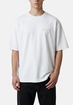 Uomo che indossa una maglietta bianca oversize semplice con testo in rilievo "4US" sul petto sinistro, abbinata a pantaloni neri, sfondo neutro.