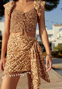 Donna in un crop top floreale marrone e beige con maniche a volant e una gonna avvolgente coordinata con dettagli in frange a conchiglia.
