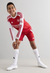 Maglietta da calcio rossa dell'Arsenal con accenti bianchi, pantaloni corti rossi, calze bianche e scarpe bianche. Presenta loghi degli sponsor e il design a tre strisce.