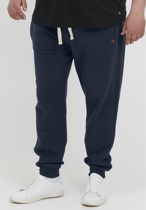 Navy blue jogger pants with cream drawstrings, side pockets, and a small brown logo patch. Soft fabric with a relaxed fit, ribbed cuffs.