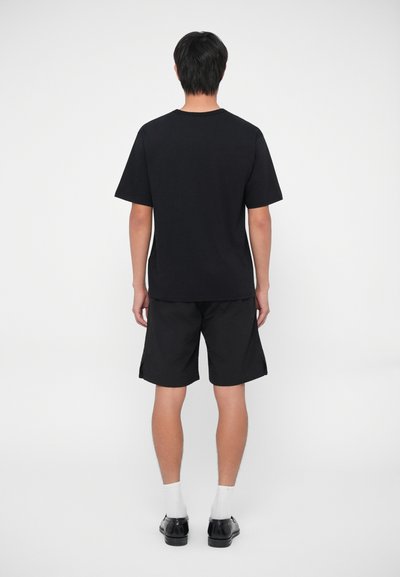 T-shirt nera a maniche corte e pantaloni corti neri, entrambi in una vestibilità comoda. La T-shirt ha un collo rotondo. Il modello indossa calze bianche e scarpe nere.