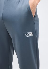 Χέρι ατόμου στην τσέπη σκούρου γκρι παντελονιού με υφή και λευκό λογότυπο The North Face στον μηρό, σε απλό φόντο.