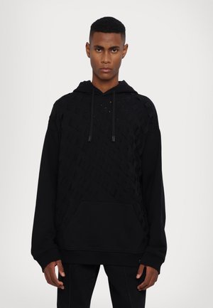 Black hoodie with woven texture on the upper half, featuring a front pocket and drawstring hood. Soft fabric with cut-out details.