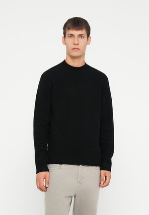 Schwarzer Strickpullover mit Rundhalsausschnitt, strukturiertem Gewebe, gerippten Bündchen und Saum, getragen über hellgrauen Hosen. Einfaches, einfarbiges Design.