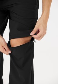 Svarta zip-off byxor har en zip-sektion vid knäet som möjliggör konvertering till shorts. Materialet verkar lätt och har en slät textur.