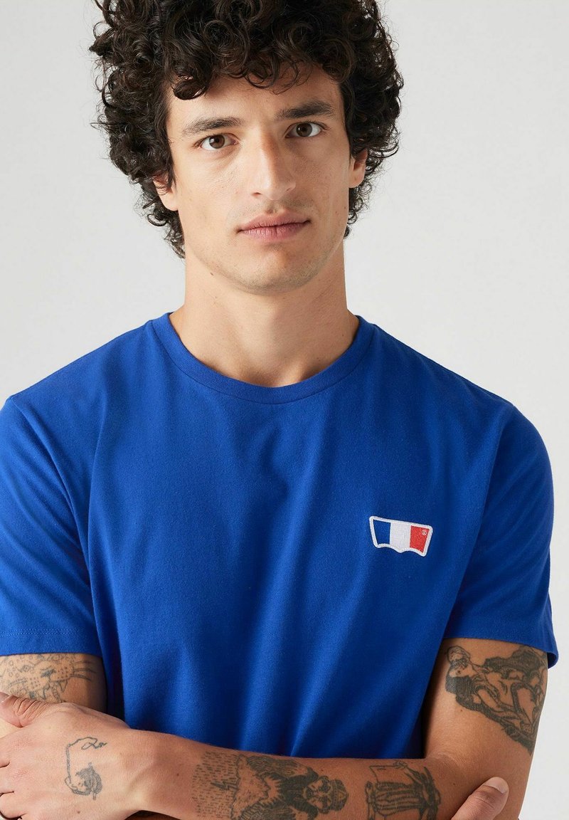 Camiseta de manga corta azul, hecha de una tela suave, con un pequeño logotipo bordado de la bandera francesa en el lado izquierdo del pecho. Textura suave.