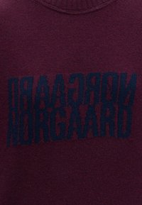 Tissu tricoté bordeaux foncé avec texte noir, verticalement miroir et inversé, lisant « NORGAAARD » en lettres majuscules grasses.