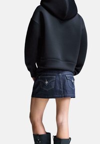 Sweat à capuche noir à texture lisse, associé à une mini-jupe en denim sombre avec poches et coutures contrastées. Bottes noires montantes jusqu'aux genoux.