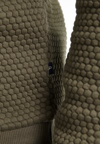 Tessuto verde oliva con texture, caratterizzato da un motivo a nido d'ape e orlo a costine. Presenta un piccolo etichetta blu scuro cucita sul lato.
