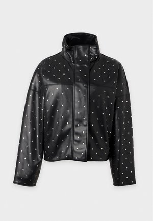 Le modèle se tient devant un fond de studio clair, vêtu d’une veste en cuir noire cloutée, d’un haut blanc, d’un pantalon anthracite et de bottes noires.