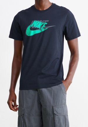 Homme portant un t-shirt Nike noir avec un logo turquoise et un short cargo gris, debout avec une main dans la poche sur un fond blanc.
