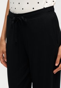 Gros plan d’un pantalon noir à taille à cordon porté avec un haut blanc à pois noirs, montrant les mains reposant sur les côtés.