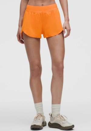 Person med slanke ben iført lyse orange atletikshorts, lyse beige sokker og beige løbesko, stående mod en ensfarvet baggrund.