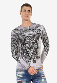 Langärmliges T-Shirt in Hellgrau mit schwarz bedruckten Grafiken, die kunstvolle Designs, Text und eine strukturierte Optik aufweisen. Kombiniert mit abgetragenen blauen Jeans.