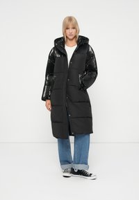 Zwarte pufferjas met een glanzend zwarte nylon bovenkant en matte onderkant, capuchon, drukknoopsluiting en twee zijzakken. Gemaakt over een blauwe spijkerbroek en zwarte sneakers.