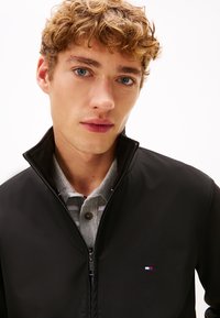 Sort lynlåsjakke med høj krave, med en lille multifarvet logo på venstre bryst, båret over en grå skjorte med krave.