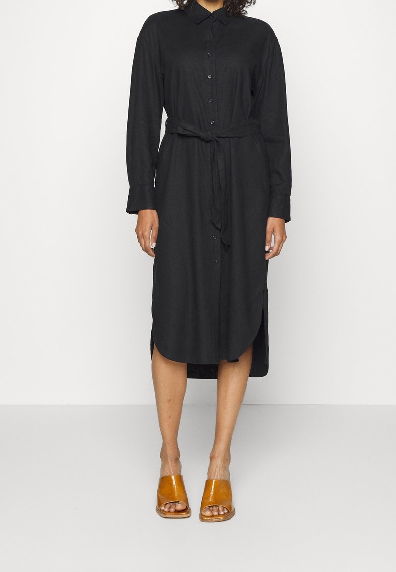 Robe chemise noire à manches longues, avec une taille ceinturée, des fentes sur les côtés et une texture lisse, associée à des sandales à talons ouverts marron.