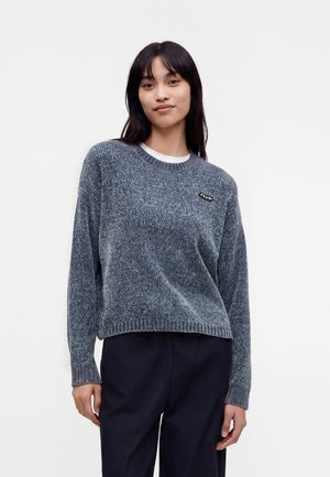 Jeune femme aux longs cheveux foncés portant un pull en maille gris avec un petit logo sur la poitrine et un pantalon noir, debout devant un fond uni.