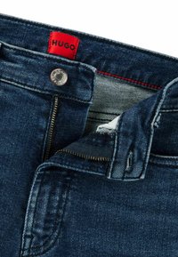 Vaqueros de mezclilla azul oscuro con una etiqueta roja marcada "HUGO". Cuenta con un cierre de cremallera de latón y un botón plateado. Ligero desgaste en la textura de la tela.