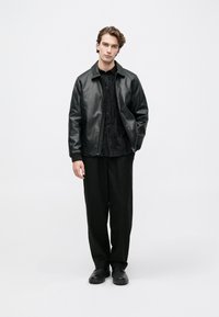 Giacca bomber in pelle nera, camicia nera con texture, pantaloni a righe e scarpe nere, il tutto presentato su uno sfondo bianco uniforme.