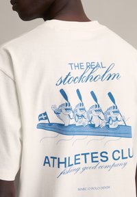 Wit katoenen T-shirt met een blauwe grafische print van cartoonvissen in een boot, met de tekst "THE REAL stockholm ATHLETES CLUB" en een vlag.