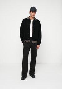 Črna pletena kardigan s sprednjimi gumbi, oblečena čez karo srajco, v kombinaciji s črnimi hlačami in prilegajočo se kapo, stoji na enobarvni podlagi.