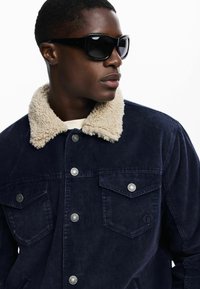 Mornarska kordura jakna s krem krznenim ovratnikom, srebrnim dugmadi na preklop i dva džepa na prsima. Nosi se s crnim naočalama.