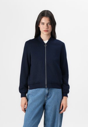 Une jeune femme se tient contre un fond blanc, portant une veste bomber bleue marine et un jean bleu clair, avec une expression neutre.