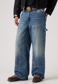 Los pantalones cargo de mezclilla azul claro presentan grandes bolsillos, un ajuste holgado, costuras amarillas y un logotipo bordado distintivo en la cintura.