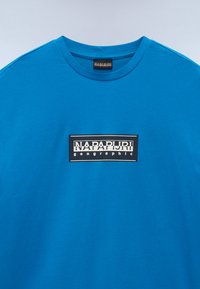Blå bomulds t-shirt med rund halsudskæring og et centralt sort rektangulært logo med hvid tekst, der står "NAPAPURI geographic."