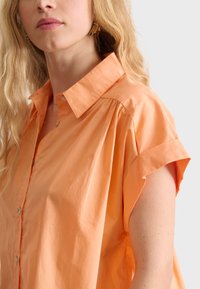 Korte mouwen oranje katoenen shirt met een button-down kraag, met subtiele plooien op de schouder en een gladde, lichte textuur.