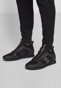 Fötter i svarta EA7 Emporio Armani-sneakers med guldfärger och svarta joggerbyxor på ett ljust grått golv.