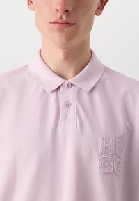 Šviesiai violetinė polo marškinėliai su klasikine apykakle, dviem mygtukais priekyje ir išsiuvinėtu "HUGO" logotipu krūtinėje. Textūruota medžiaga.