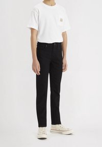 Persona vestida con una camiseta blanca de manga corta y bolsillo en el pecho, jeans negros y zapatillas de lona color crema, de pie frente a un fondo blanco.