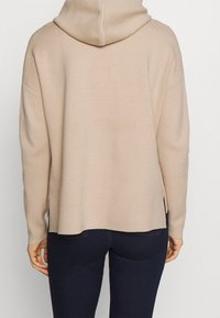 Beiger Hoodie mit lockerer Passform, ausgestattet mit Kapuze, langen Ärmeln und seitlichen Schlitzen am Saum. Weicher Stoff und minimalistisches Design.