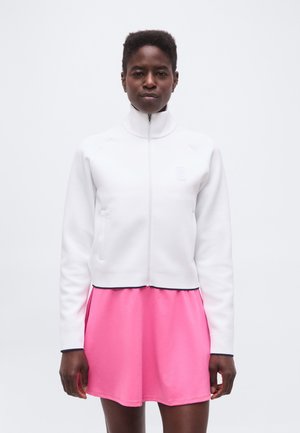 Άτομο με κοντά μαλλιά που φοράει λευκό μπουφάν με φερμουάρ και φωτεινή ροζ φούστα, στέκεται μπροστά σε απλό λευκό φόντο.