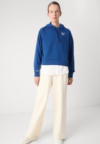 Felpa blu cropped con cappuccio e cordino, dettaglio del logo, indossata sopra una camicia bianca, abbinata a pantaloni a gamba larga color crema e sneakers bianche.