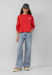 Rode cropped sweatshirt met een klein logo op de borst, gecombineerd met lichtblauwe wijde jeans en crèmekleurige plateau-sandalen. Een grijze pet accentueert de outfit.