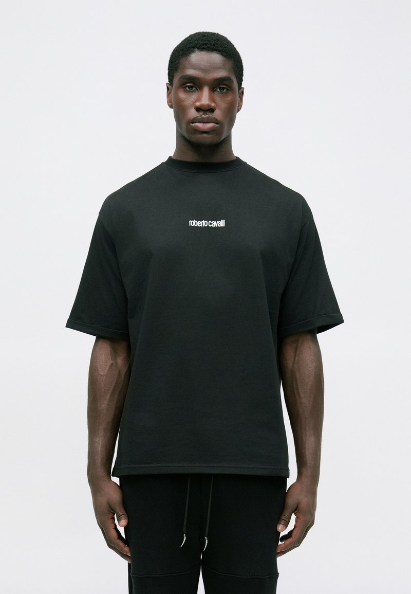 Homme portant un t-shirt oversized noir avec le logo blanc "roberto cavalli" centré sur la poitrine, associé à un pantalon noir, se tenant devant un fond uni.