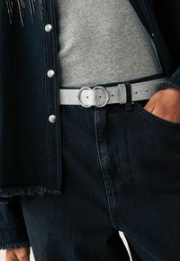 Cintura in finta pelle argentata con fibbia ovale, indossata sopra una maglietta grigia e jeans in denim scuro, con orlo sfrangiato e lavaggio scuro.