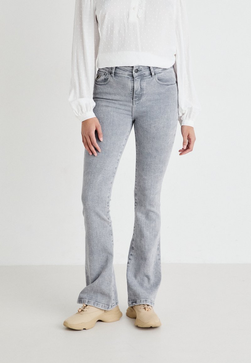 Jeans grigi flared con lavaggio chiaro, tessuto misto cotone, design a cinque tasche e cuciture visibili, abbinati a una blusa bianca con texture e scarpe beige.