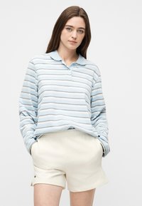 Νεαρή γυναίκα που φοράει μια μακρυμάνικη μπλούζα πόλο με γαλάζιες και λευκές ρίγες και κρεμ σορτς, με τα χέρια στις τσέπες.
