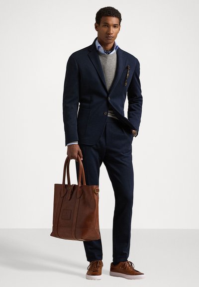 Navy suit with a fitted design, gray sweater, and light blue shirt. Brown leather tote and sneakers with textured accents.
