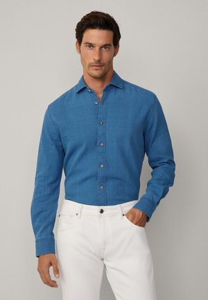 Heren overhemd met lange mouwen en knoopsluiting in middelblauwe denim. Voorzien van een kraag, vijf knopen aan de voorkant en een relaxte pasvorm. Gecombineerd met witte broeken.