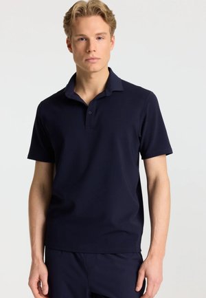 Ung mand med blondt hår står med fronten mod foran iført en ensfarvet mørk navy kortærmet poloshirt og matchende bukser.