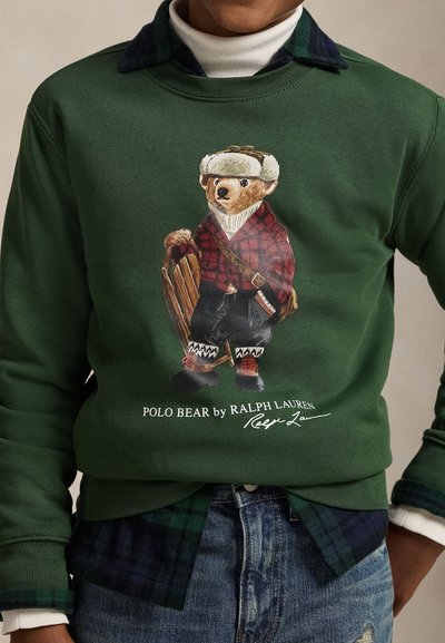 Grön sweatshirt med en bild av en björn i rutigt mönster med bruna och svarta detaljer, ovanpå en blå rutig skjorta och jeans.