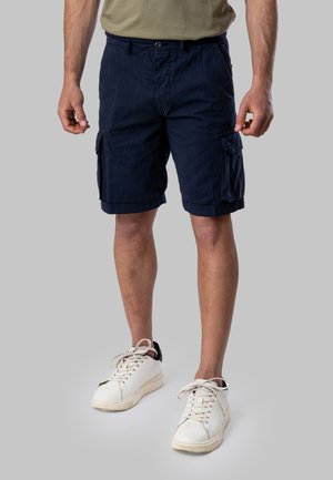 Uomo con pantaloncini cargo blu navy, t-shirt beige e sneakers bianche, in piedi davanti a uno sfondo grigio semplice.