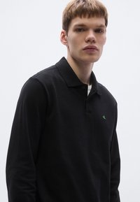 Must pika varrukaga polo-särk tekstureeritud kangaga ja rohelise logoga rinnal, klassikalise krae ja nööbitava plaketiga.