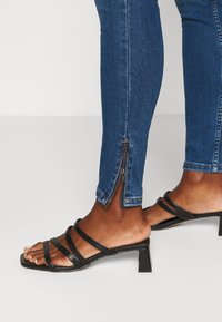 Sandalias negras con tiras y tacón medio en bloque, combinadas con jeans ajustados azules que tienen un detalle de cremallera en el tobillo. Textura suave.