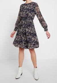 Navy floral dress with long, sheer sleeves, cinched waist, and flared skirt; features cream and peach floral patterns. Paired with white ankle boots.